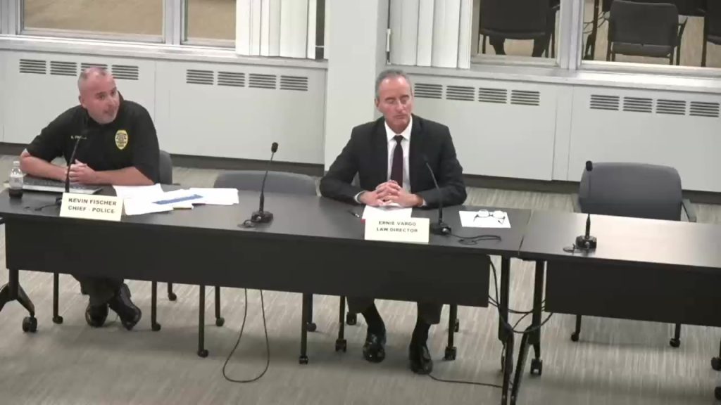 Police Chief Kevin Fischer speaks into a microphone at a table beside Law Director Ernie Vargo
